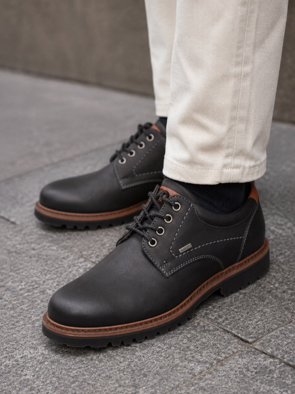 A close-up of a person wearing black leather lace-up shoes with brown soles, paired with off-white trousers and dark socks, standing on a gray stone pavement.