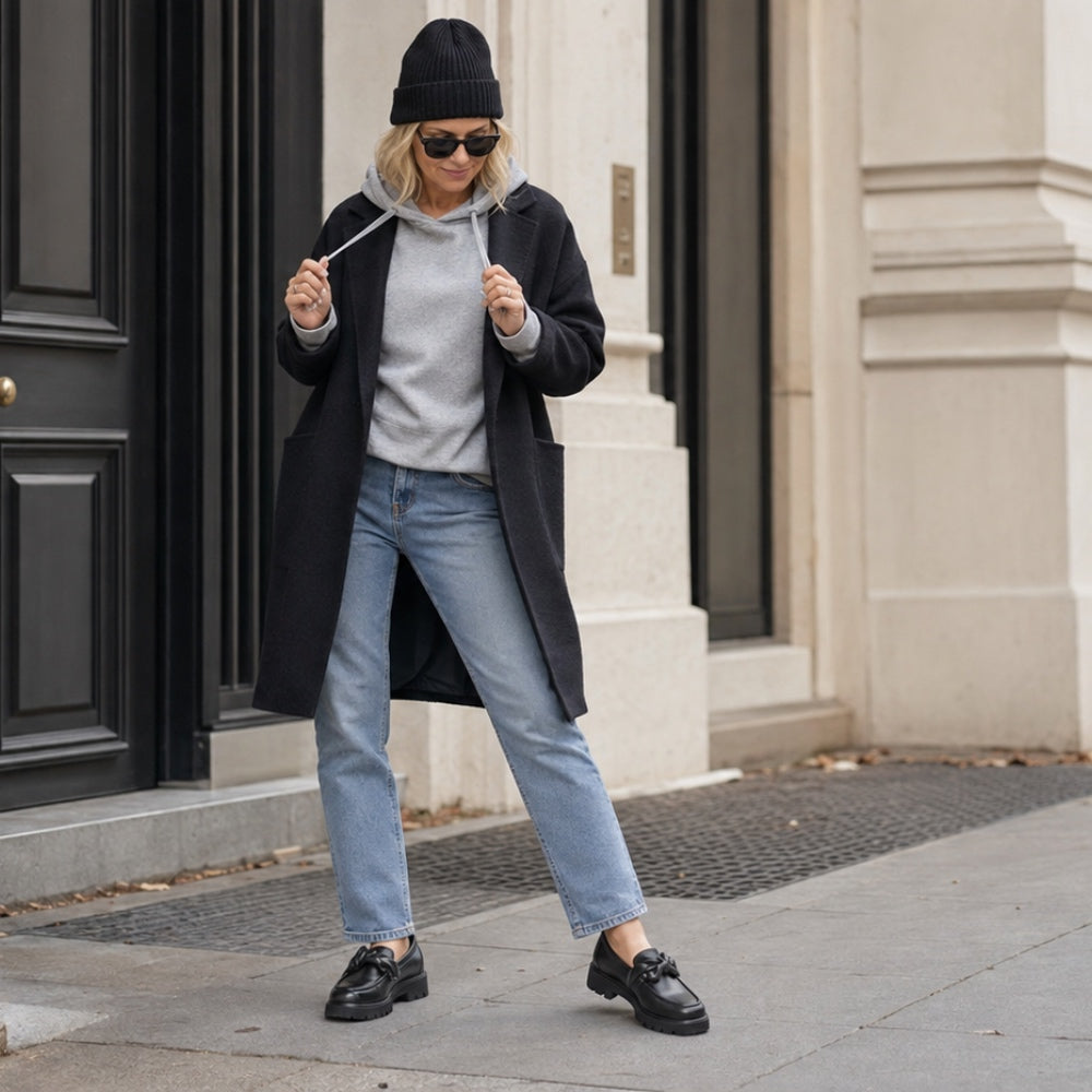A woman stands on a city sidewalk wearing a black coat, light gray hoodie, light blue jeans, black loafers, and a black beanie.