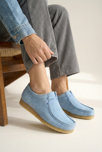 Close up of woman sitting on a wooden chair wearing a denim jacket, gray pants, and grasshopper loafers/ moccasin in baby blue