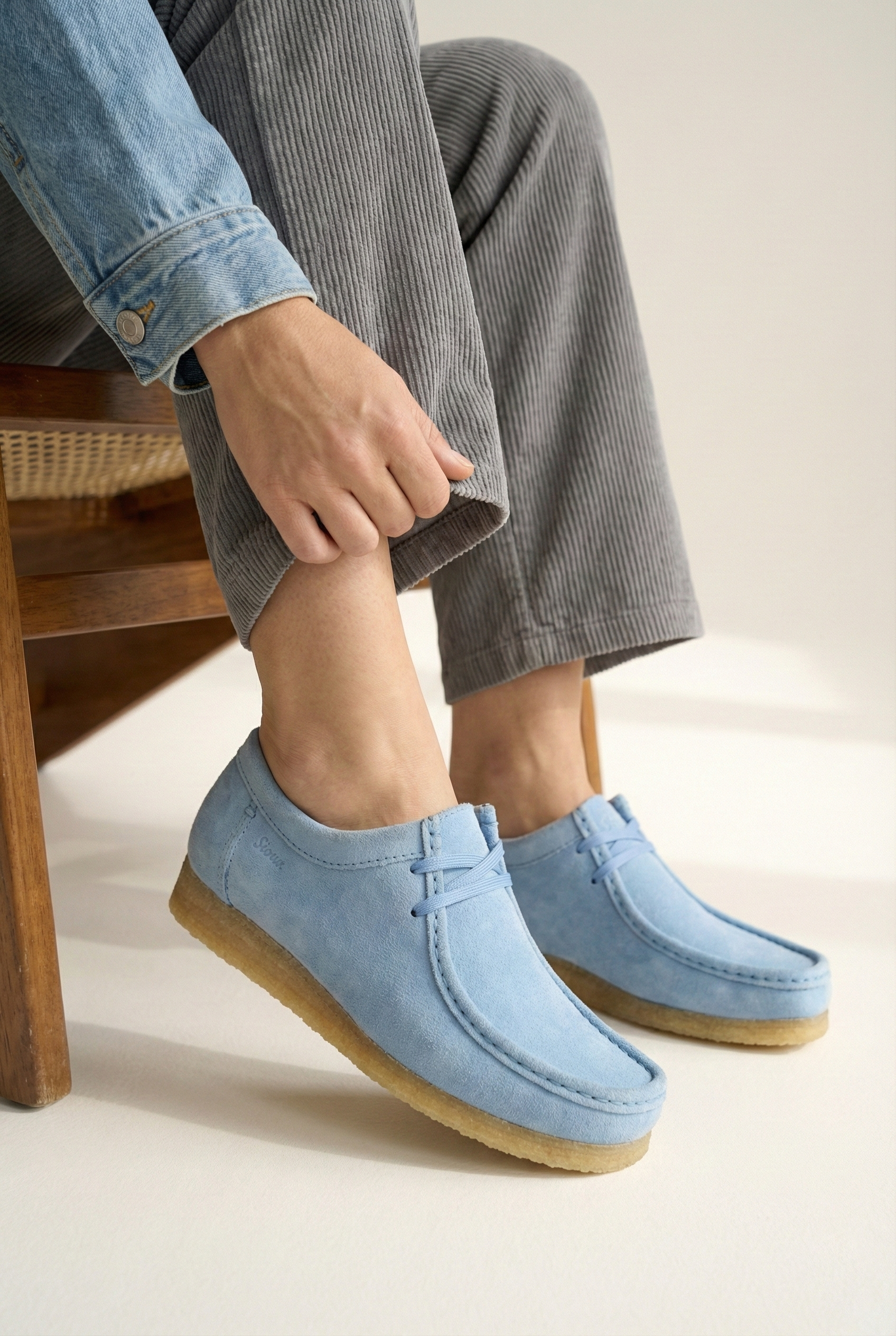 Close up of woman sitting on a wooden chair wearing a denim jacket, gray pants, and grasshopper loafers/ moccasin in baby blue